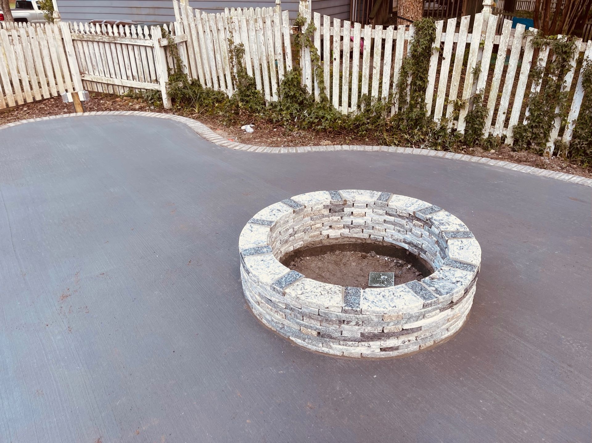 A stone fire pit in a paved area, surrounded by a white picket fence and greenery.