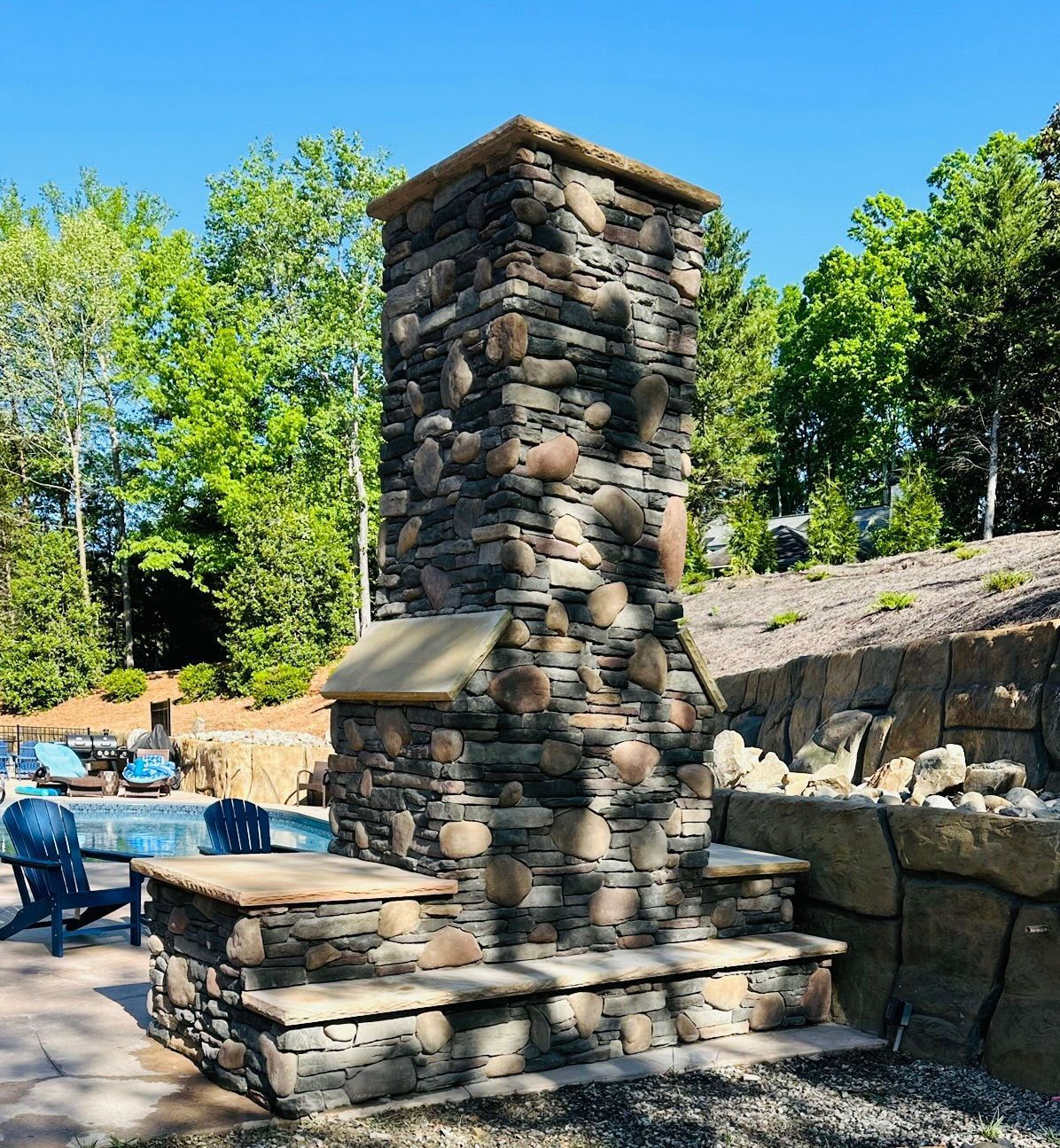 Stone outdoor fireplace with steps, pool and trees in background.
