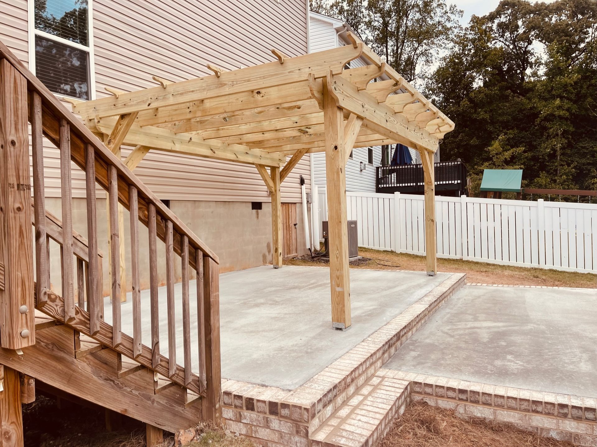 Wooden pergola over a concrete patio with stairs leading down. White fence and trees in the background.