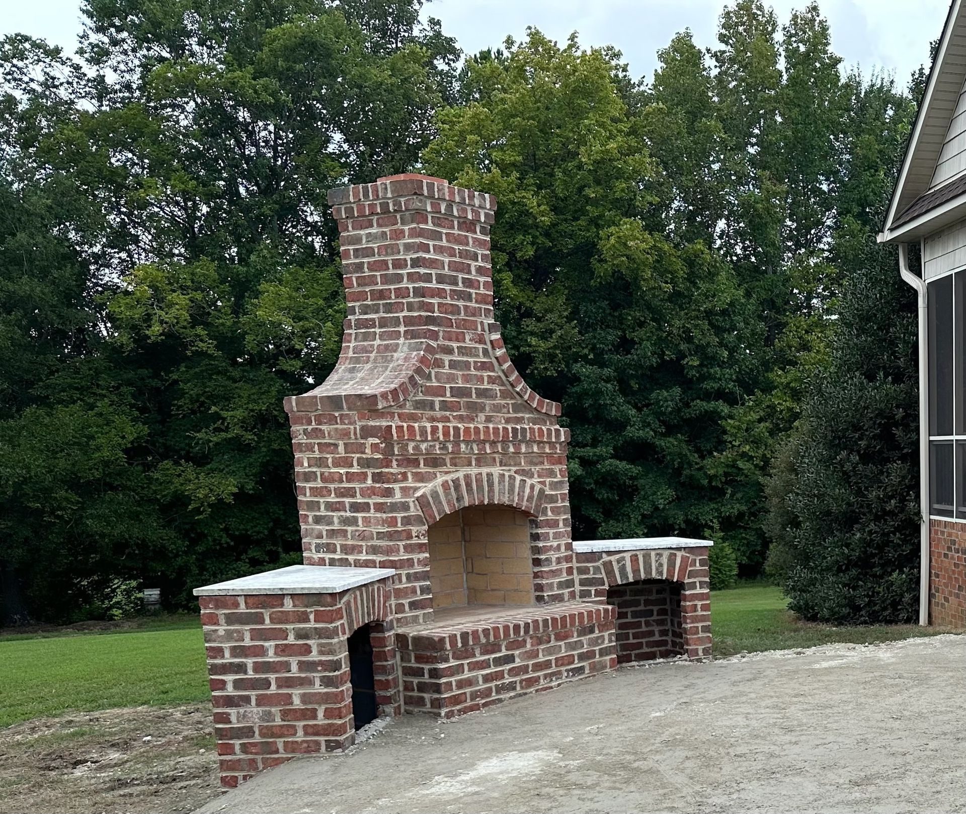 Brick outdoor fireplace with seating, in a grassy yard with trees.