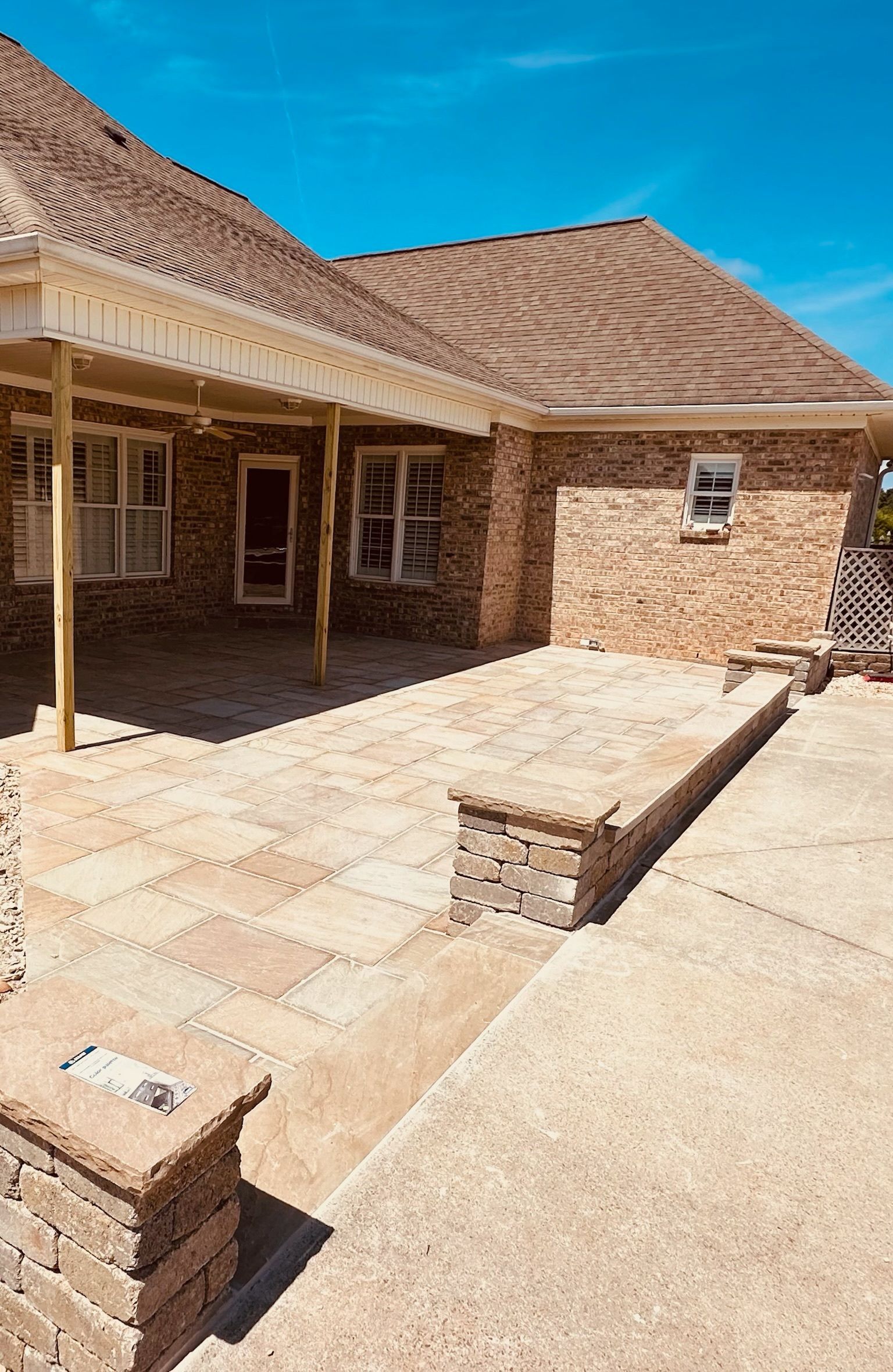 Patio with brick wall, tan pavers, and brown roof; sunny day.