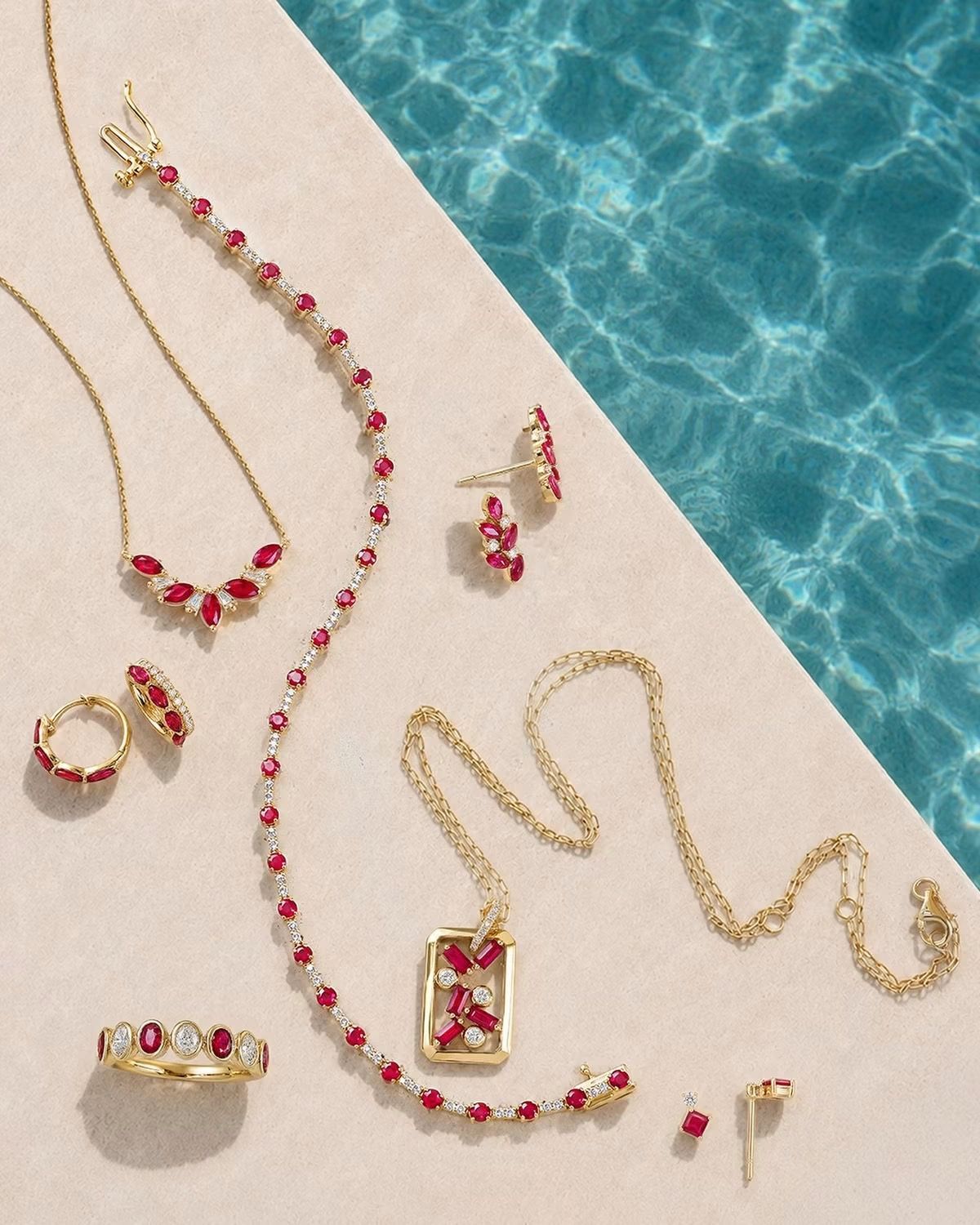 Gold jewelry with pink gemstones arranged on a beige surface beside turquoise water.