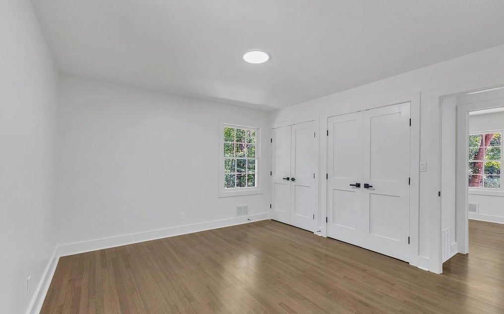 Empty room with light wood floors, white walls, and a recessed ceiling light. Two white closet doors and a window.