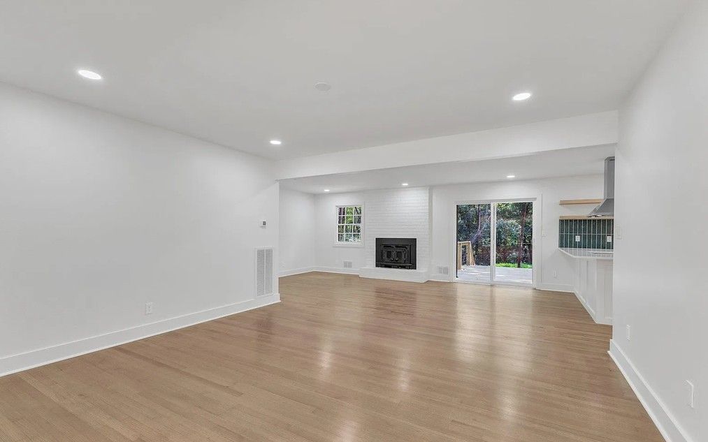 Empty, bright living area with hardwood floors, white walls, and a fireplace. Sliding glass doors lead to a backyard.