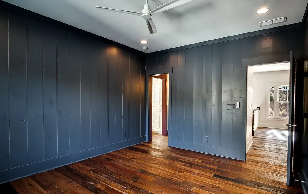 Empty room with dark blue wood panel walls, hardwood floor, and doorway to a lit room.