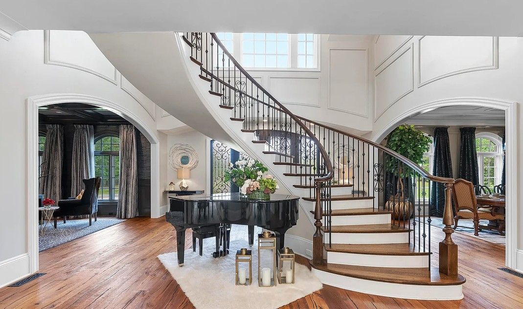Elegant foyer with curved staircase, grand piano, and arched doorways. Wooden floors and black railings.