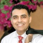 Man in a white shirt and patterned tie smiling, holding a jacket over his shoulder, with pink flowers in the background.