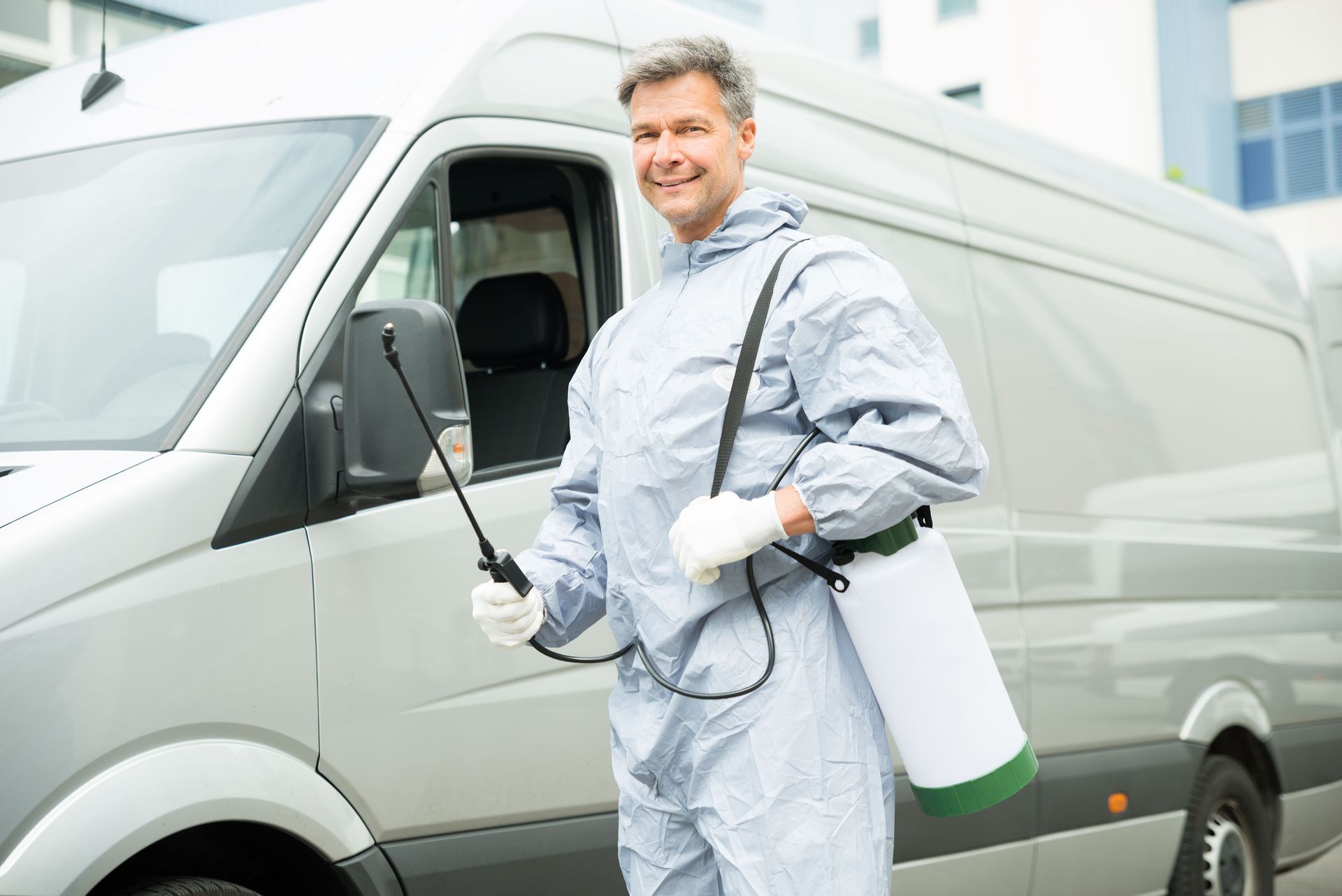 Man in protective suit with sprayer, standing next to a silver van; smiling.