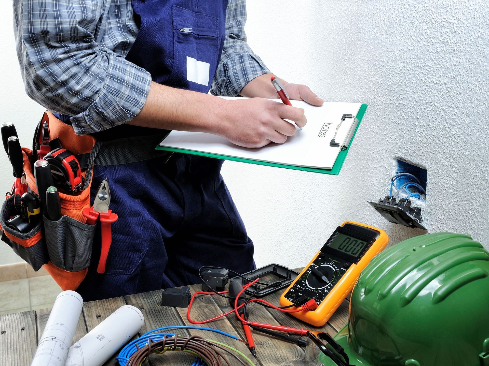 Electrician in blue overalls, taking notes on a clipboard, tools and equipment on a table.