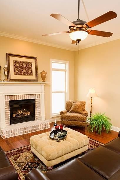 Cozy living room with fireplace, armchair, ottoman, and ceiling fan; warm tones.