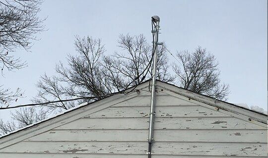White house gable with a metal antenna extending upward. Bare trees and cloudy sky.