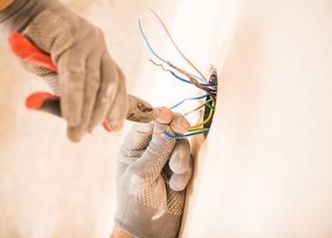 Person in gloves using pliers to work on electrical wires inside a wall.