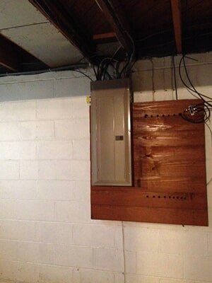 Electrical panel mounted on a wooden board on a white brick wall in a basement.