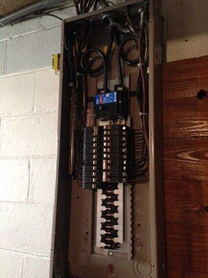 Electrical panel with numerous circuit breakers, wires, and a blue main breaker, mounted on a brick and wood wall.