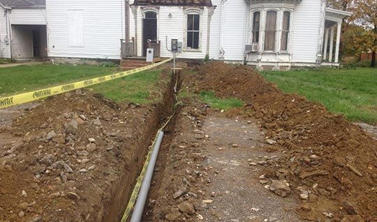 Trench dug in front of a white house with a PVC pipe inside; caution tape along edge.