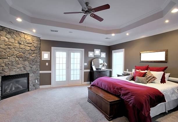 Spacious bedroom with stone fireplace, burgundy bedding, and double doors; neutral walls, tray ceiling, and ceiling fan.