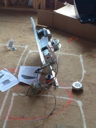 Metal appliance parts on a wooden floor with construction markings, tape measure, and paper instructions.