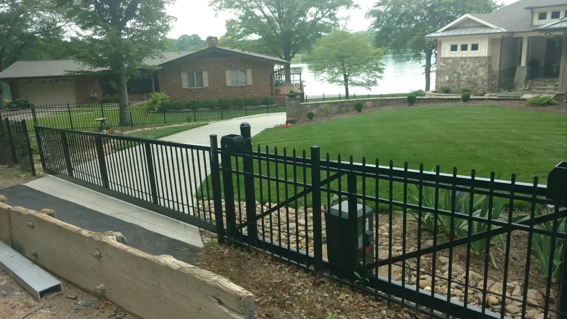 Black metal fence and gate bordering a green lawn, driveway, and residential properties.