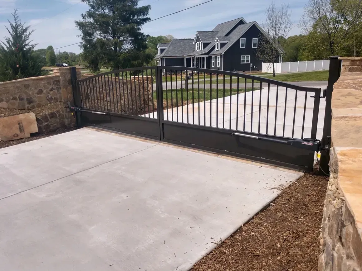 Black metal driveway gate, concrete driveway, and stone walls leading to a dark house.