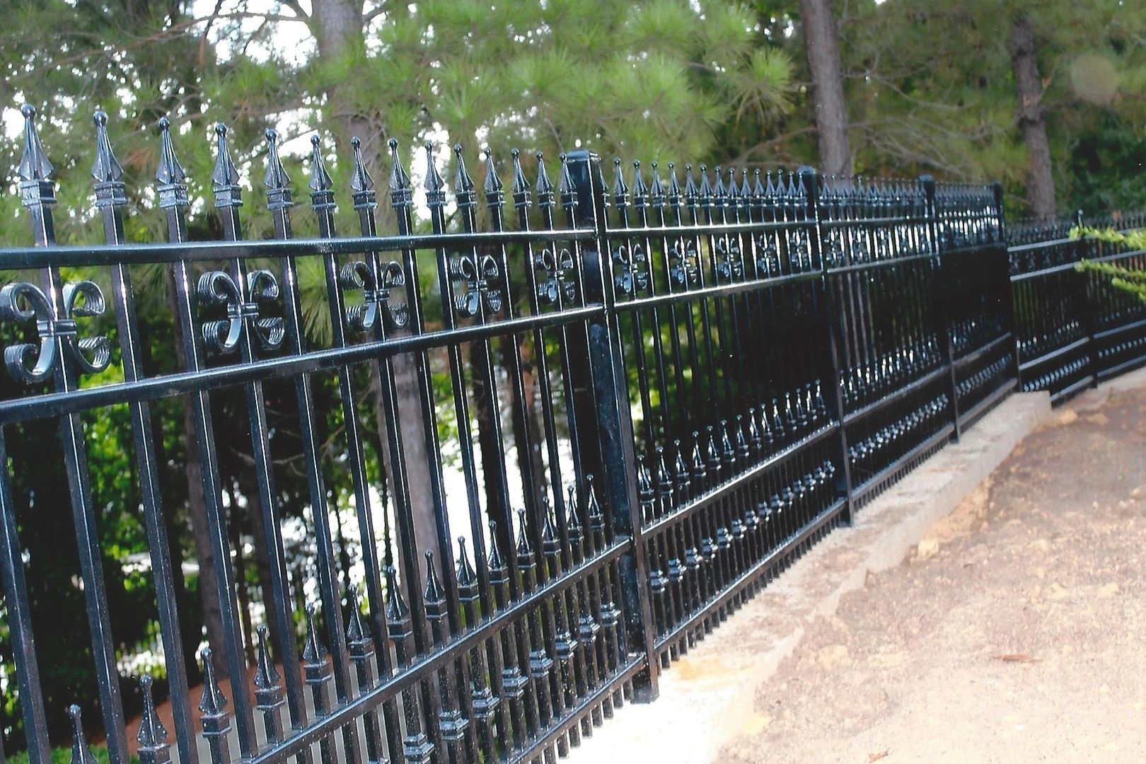 Black wrought-iron fence along a path, with ornate fleur-de-lis and finial designs, next to trees.