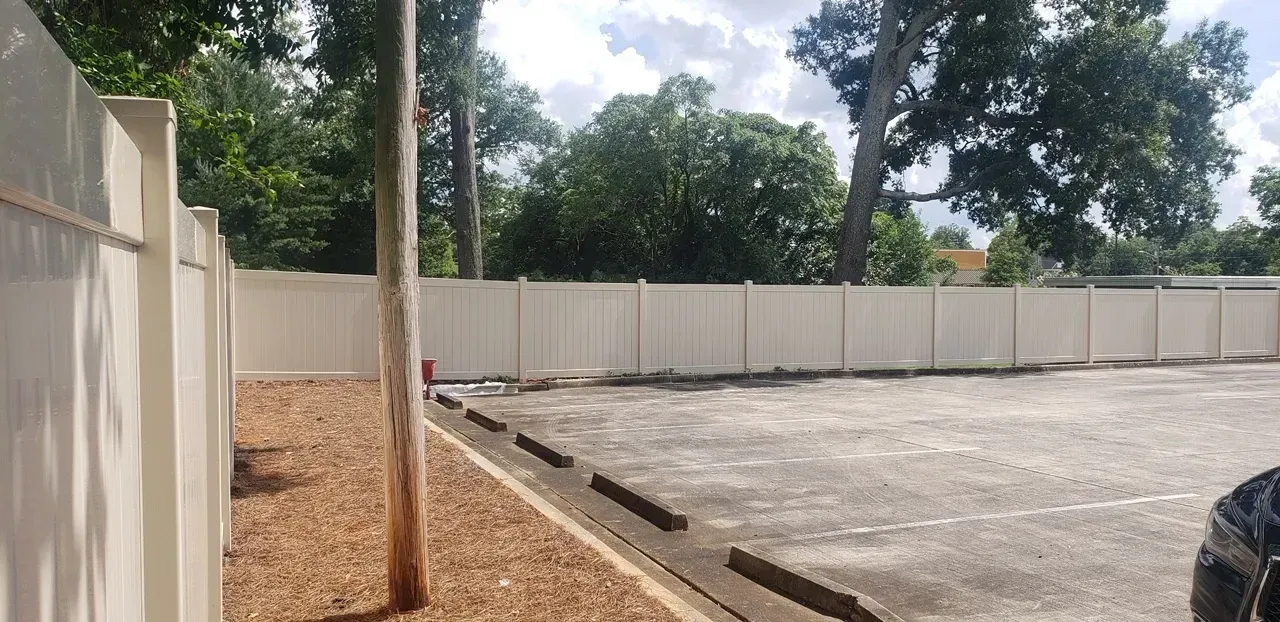 A concrete parking lot bordered by a white fence and trees. A lone tree trunk is in the middle.