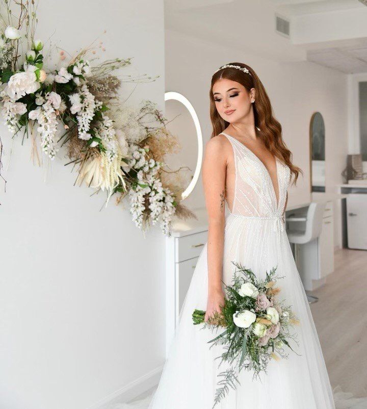 Bride in white gown holding bouquet beside floral wall arrangement in a bright room