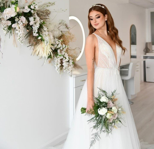 Bride in white gown holding bouquet beside floral wall arrangement in a bright room