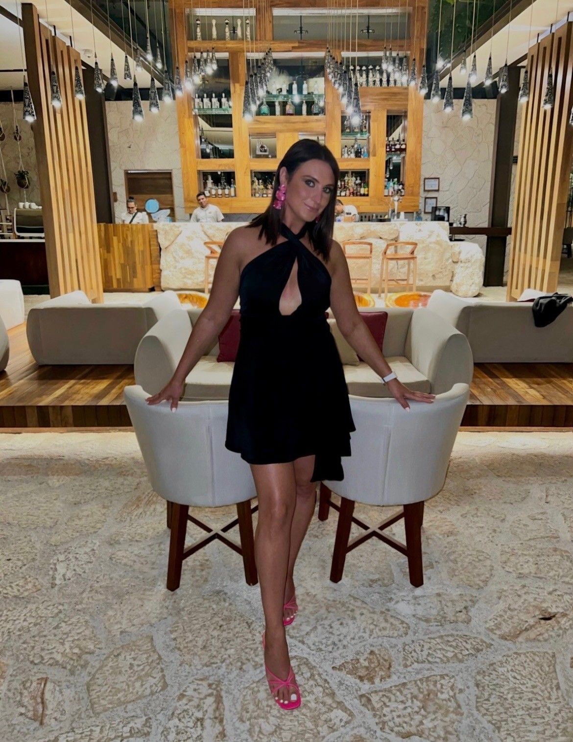 Woman in black dress and pink shoes stands between two white chairs.