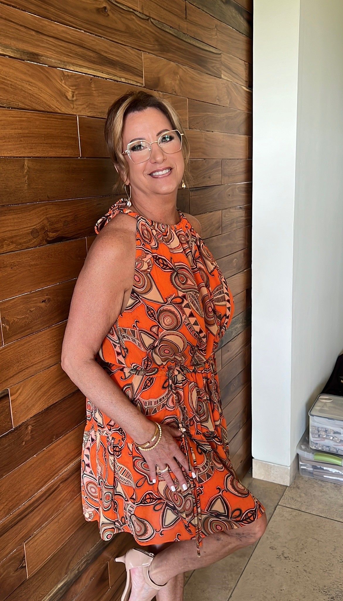 Woman in an orange floral dress and glasses leans against a wood-paneled wall, smiling.