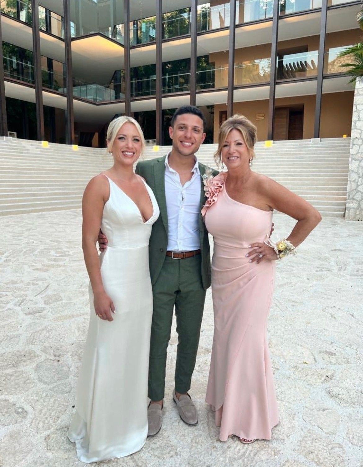Three people posing outdoors. Woman in white dress, man in green suit, and woman in pink dress smiling near a building.