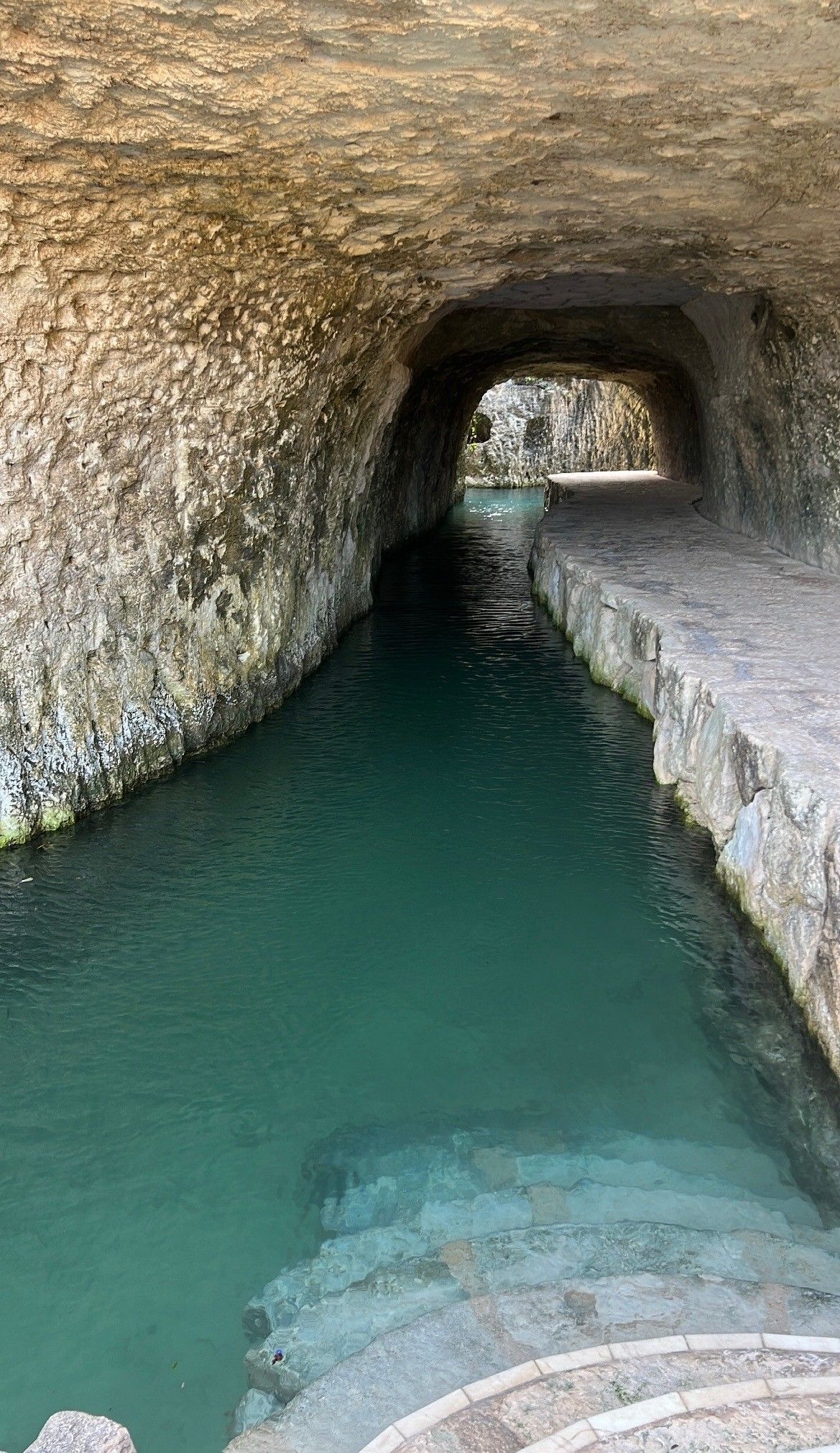 Tunnel with turquoise water and stone steps. Sunlight streams in from the opening.