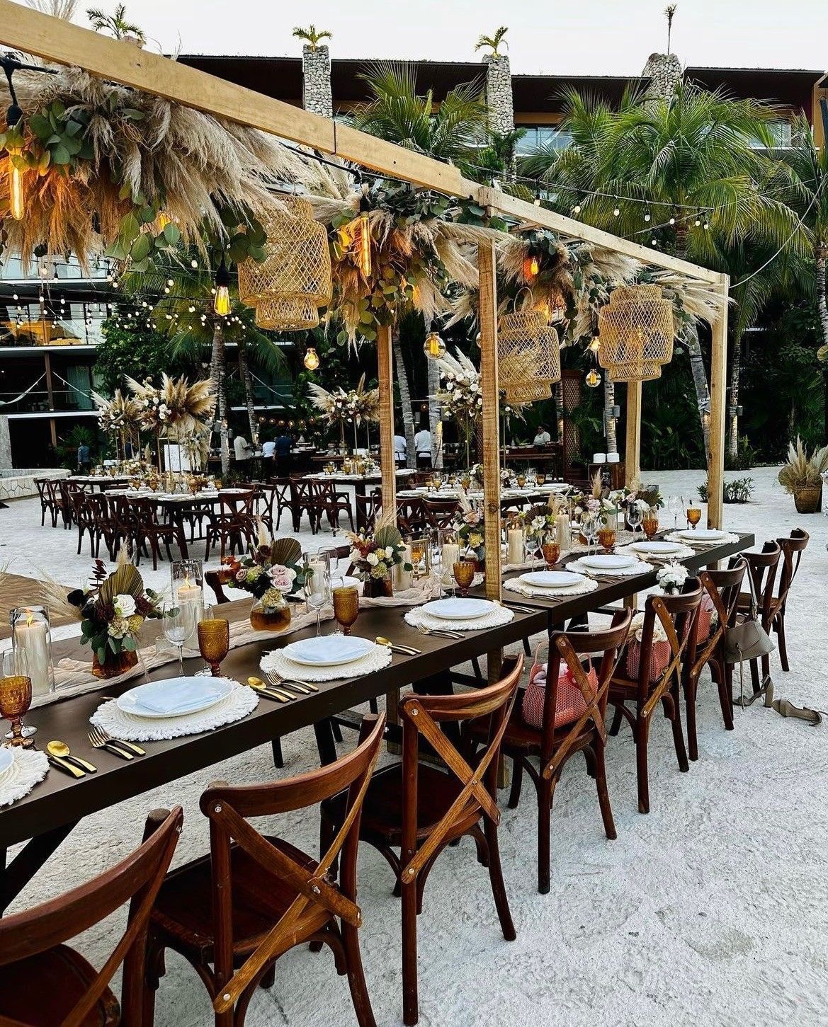 Outdoor wedding reception setup with long tables, cross-back chairs, and floral decorations.