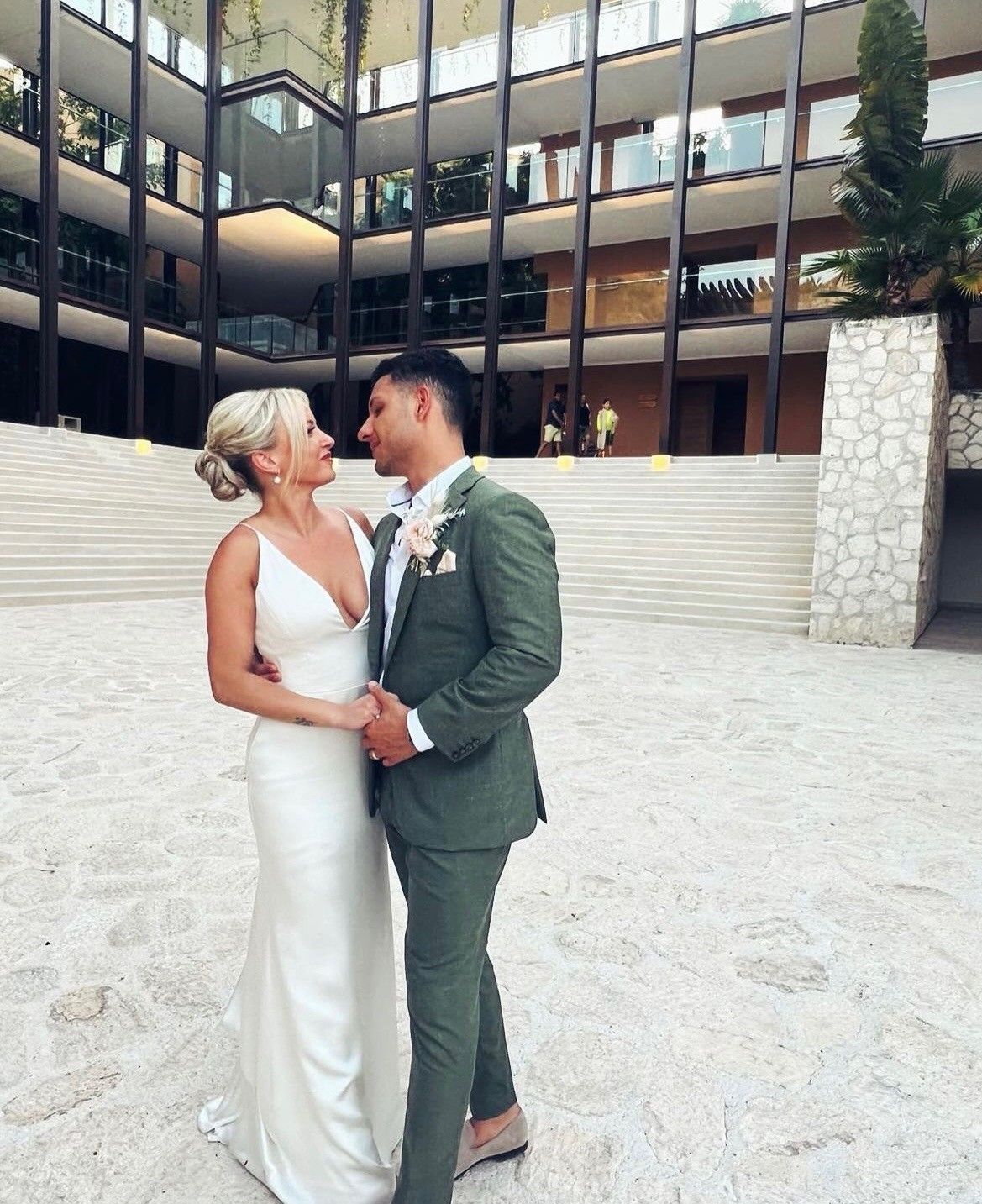 Bride and groom in wedding attire gaze at each other. Man in green suit, woman in white dress, in front of a modern building.