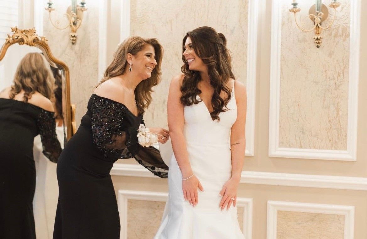 Bride in white dress smiles at woman helping her; another woman in black dress nearby.