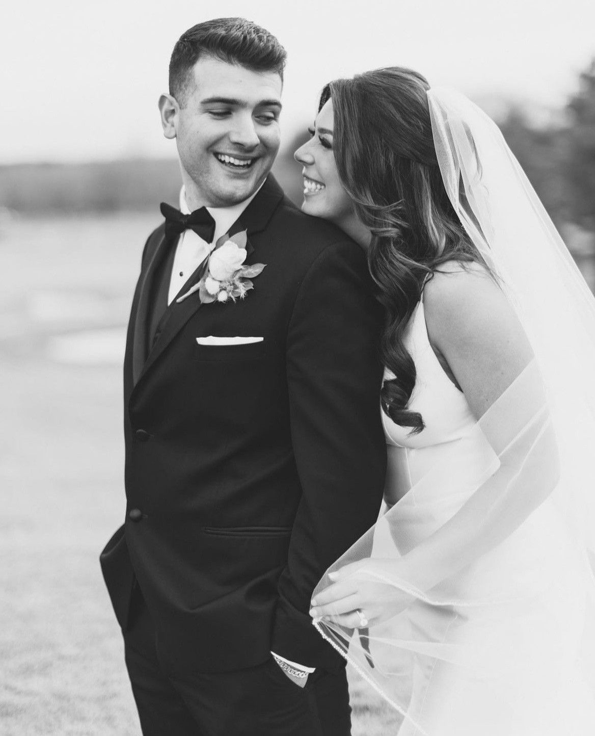 Bride and groom smiling at each other. Bride wearing a veil and holding groom's hand. Formal wear. Outdoor setting.