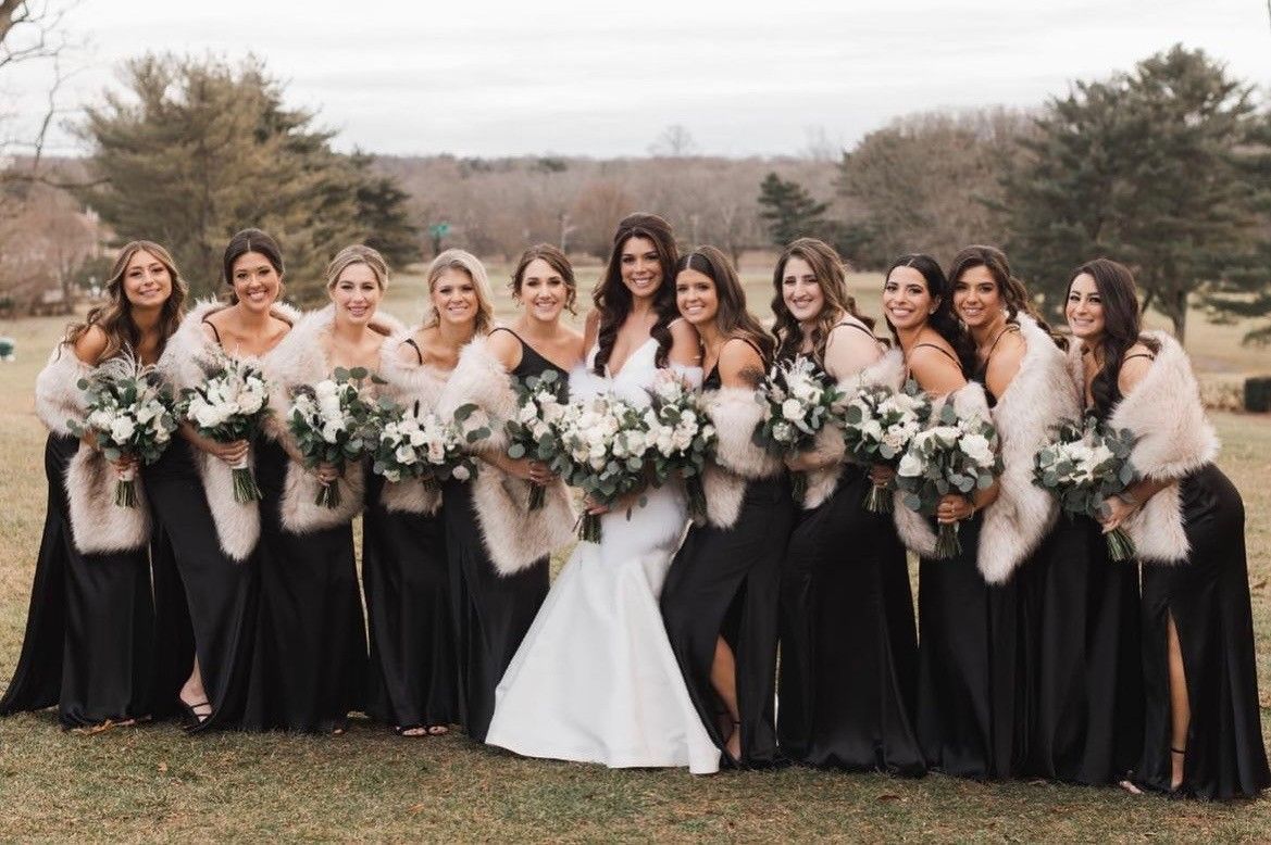 Bride and bridesmaids in black gowns and faux fur shawls, holding bouquets outdoors.