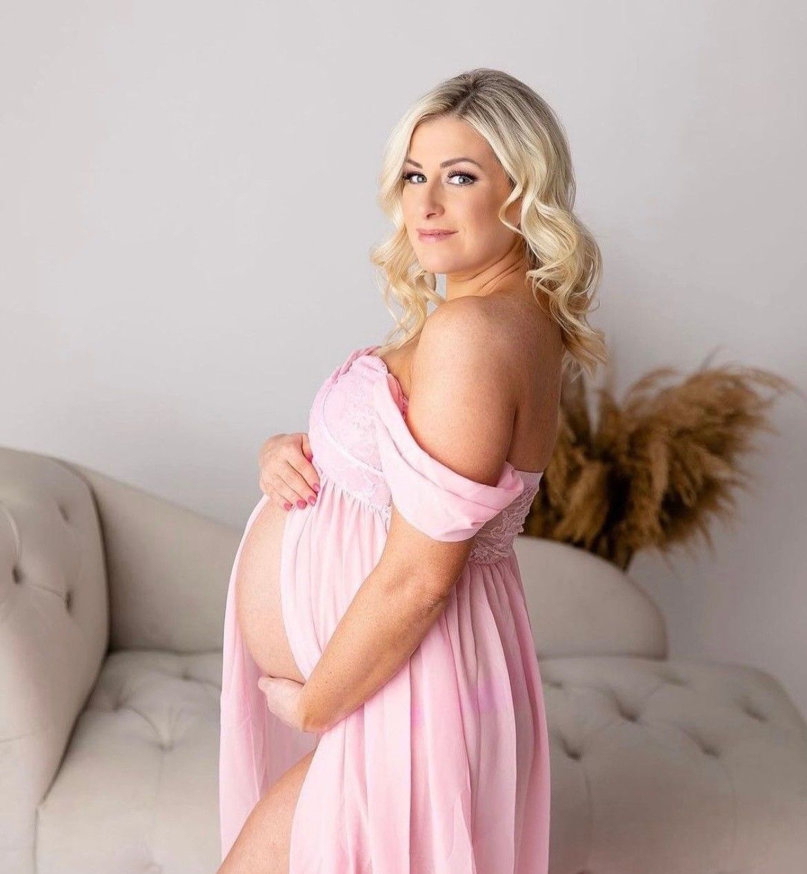 Pregnant woman in a pink dress, hand on belly, posing in front of a white couch, neutral expression.
