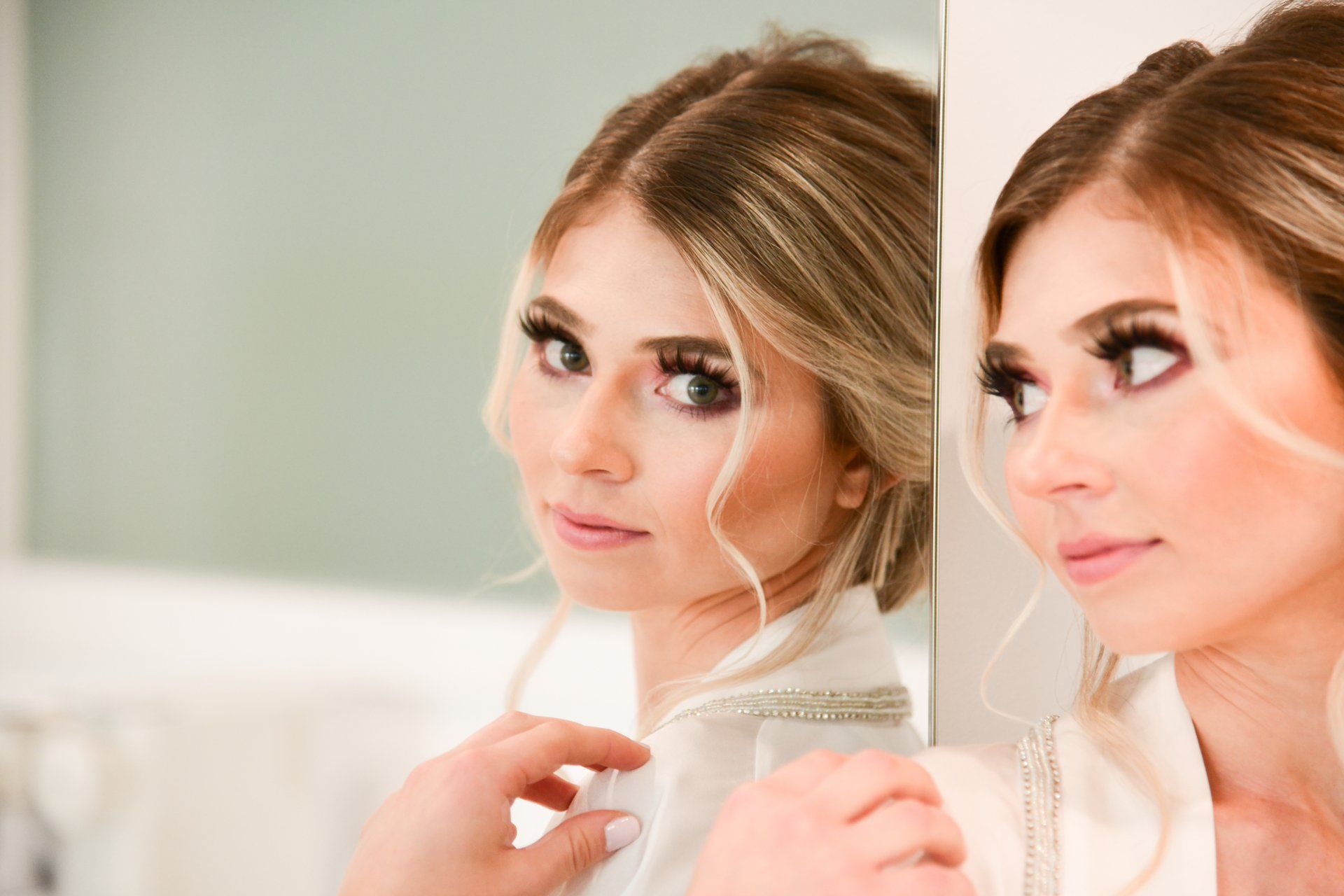 Blonde woman in a white robe looking at her reflection in a mirror; soft lighting.