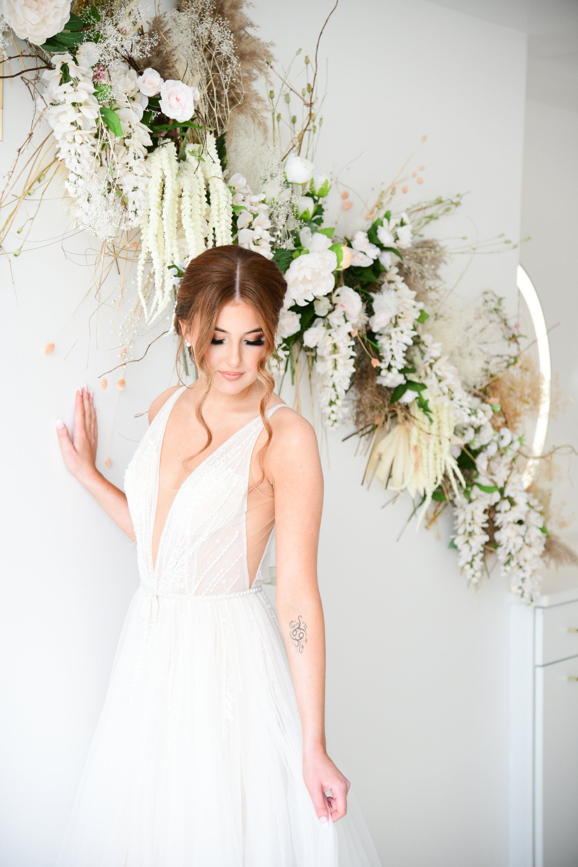 Bride in white dress, leaning against wall with floral arch.