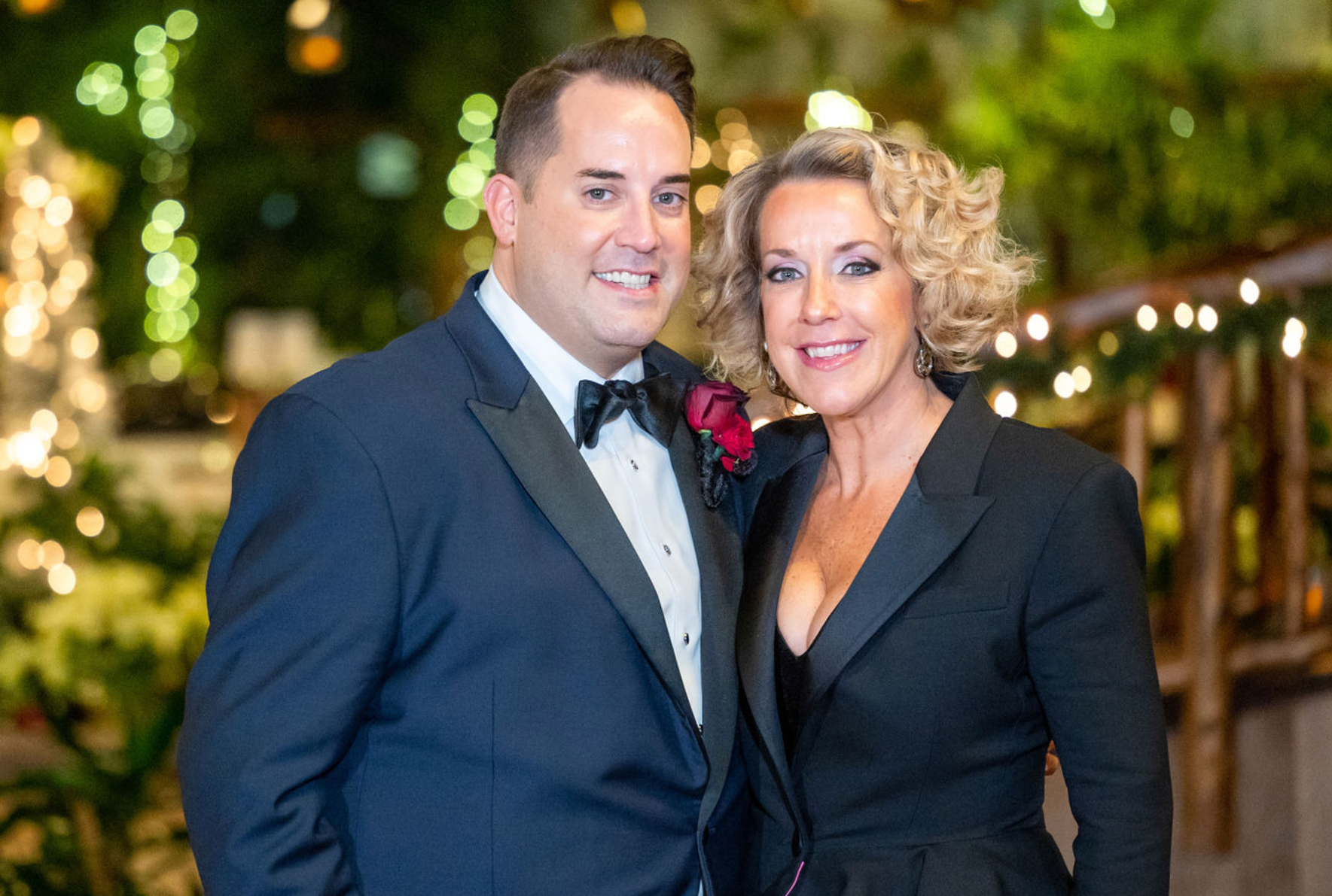 Man in tuxedo and woman in black blazer pose, smiling near string lights.