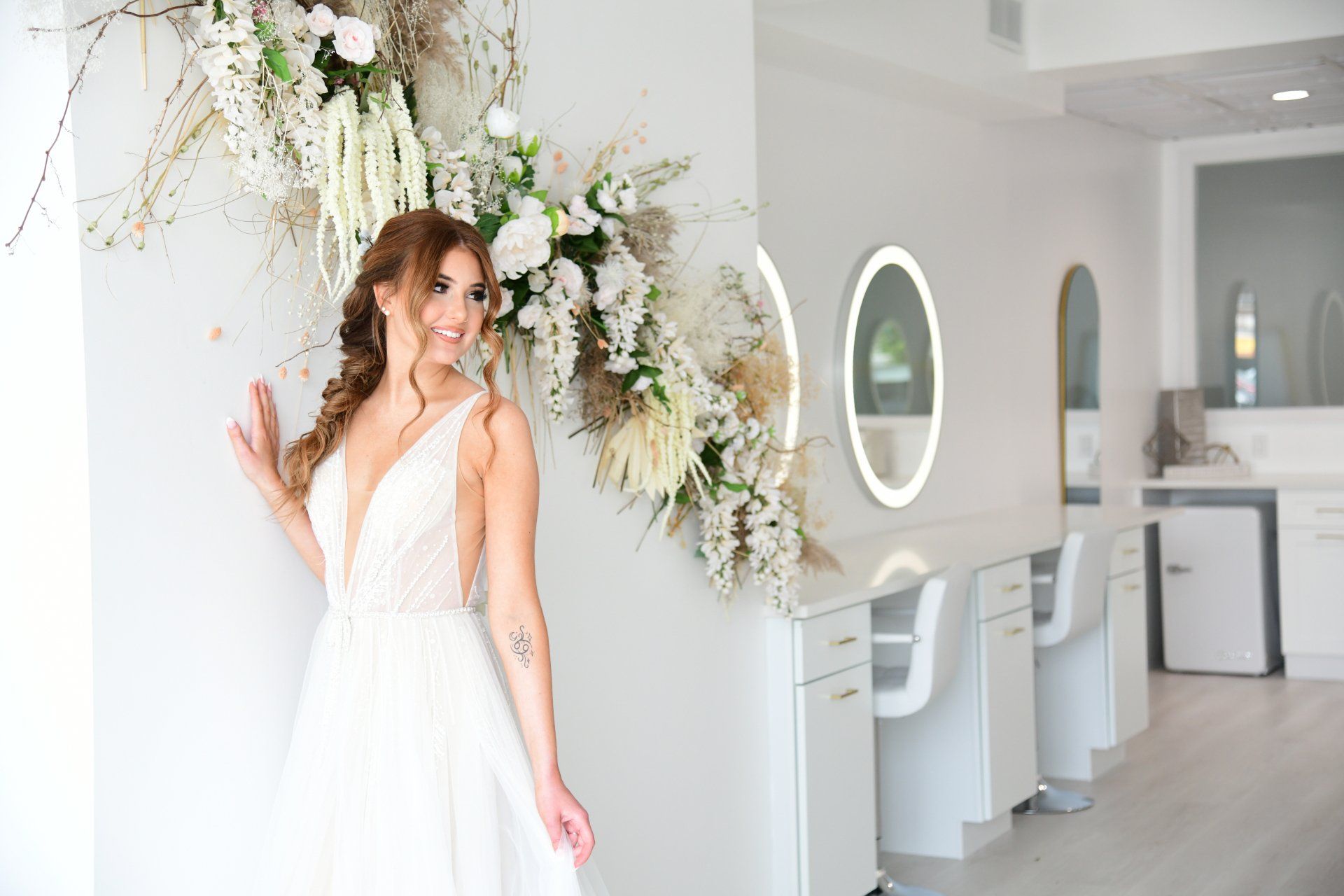 Woman in wedding dress smiles near floral arrangement in a white salon.