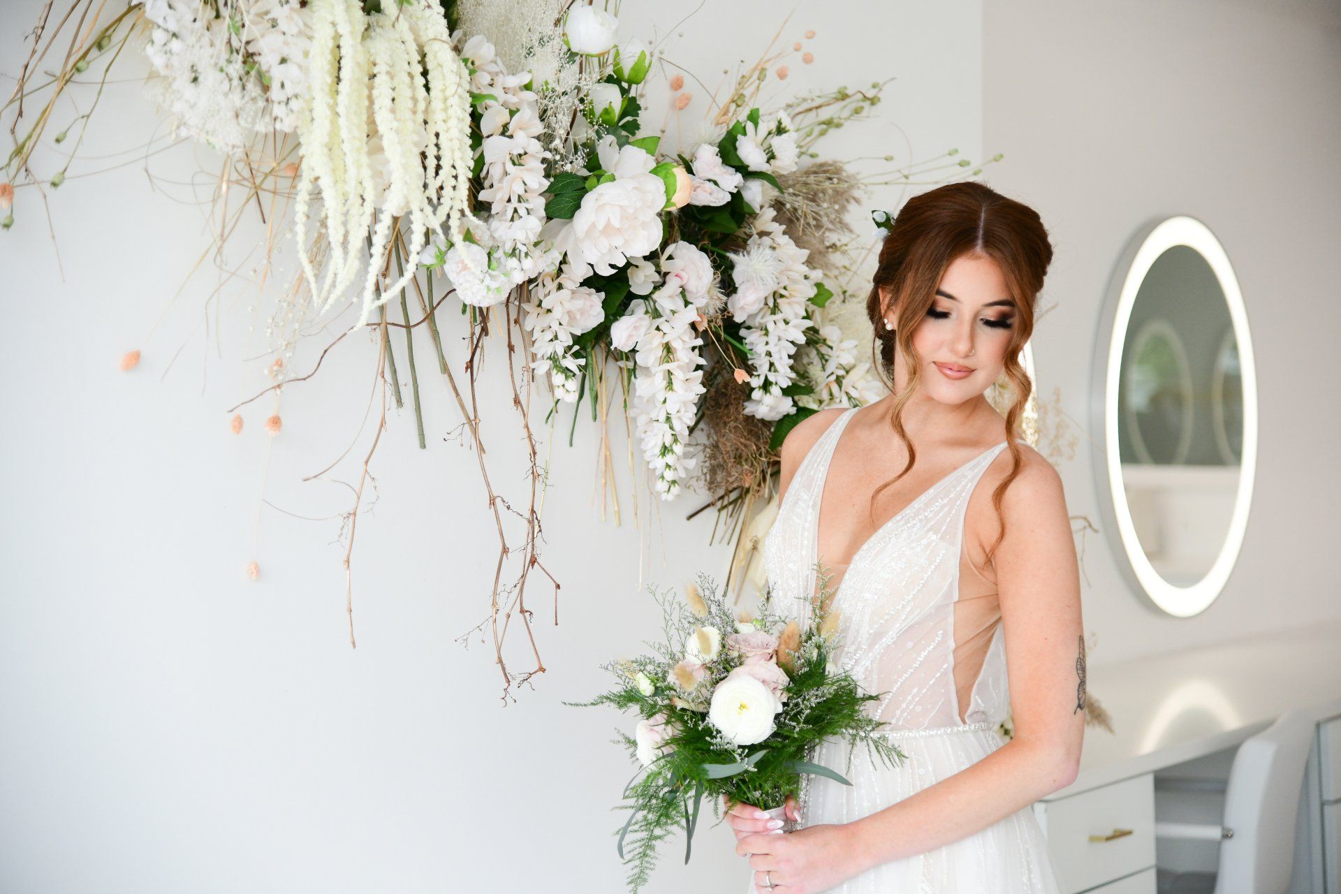 Bride in white dress, holding bouquet, standing next to floral wall decor. Bright, airy room.