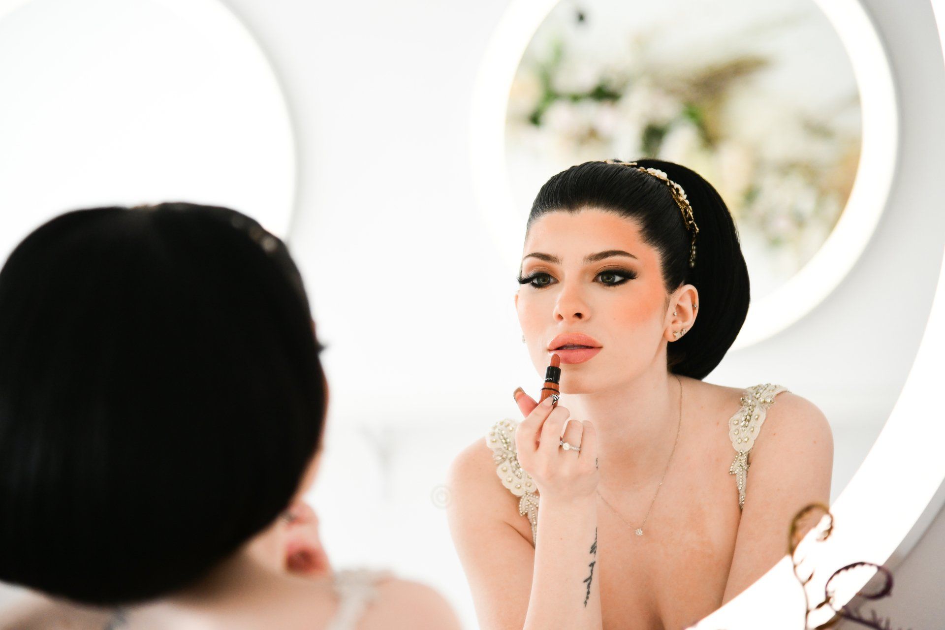 Woman applying lipstick in a mirror, wearing a cream-colored gown and a high bun in a white-walled room.