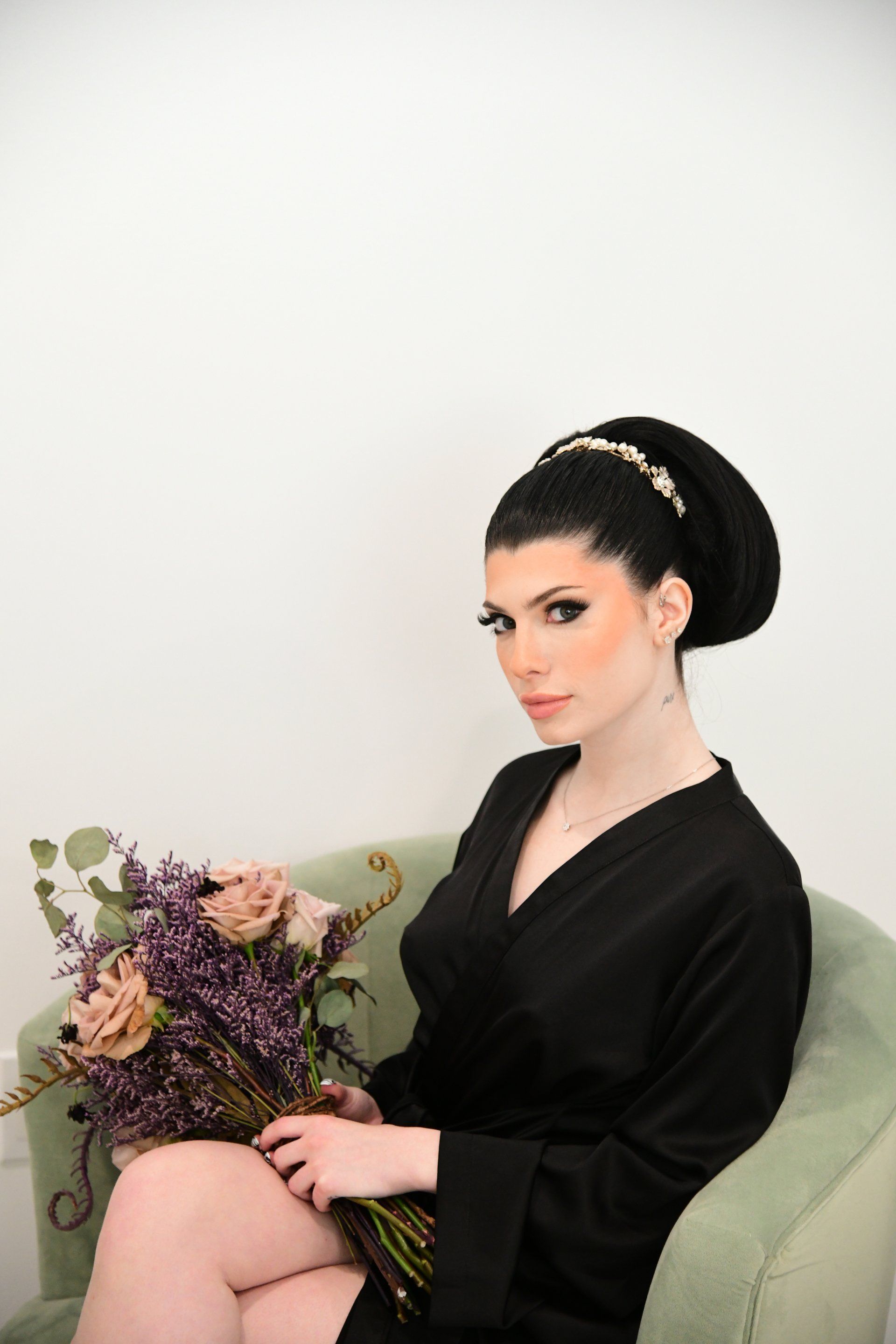 Woman in black robe holding flowers, sitting in an armchair with updo, and decorated headband.