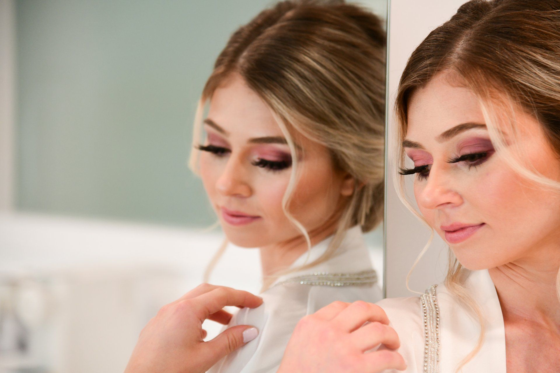 Woman with light makeup, looking down, reflected in a mirror. Wearing a white robe.