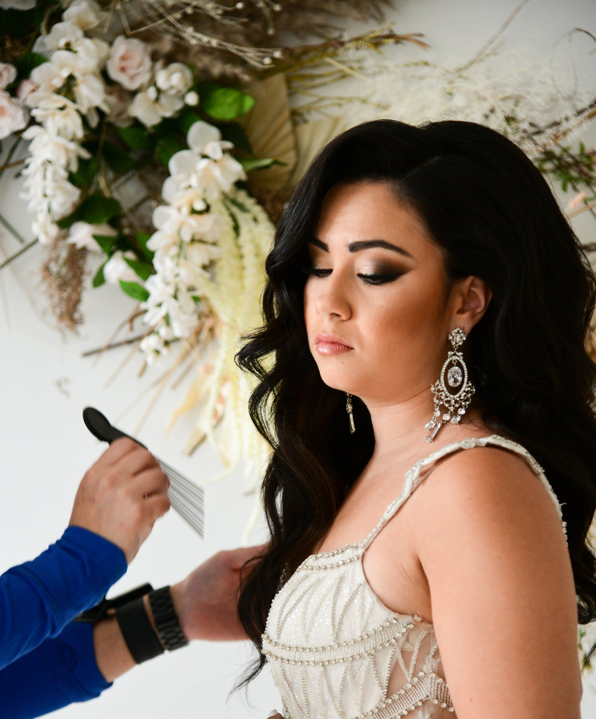 Woman in formal dress with long, wavy dark hair, wearing large earrings, having makeup adjusted.