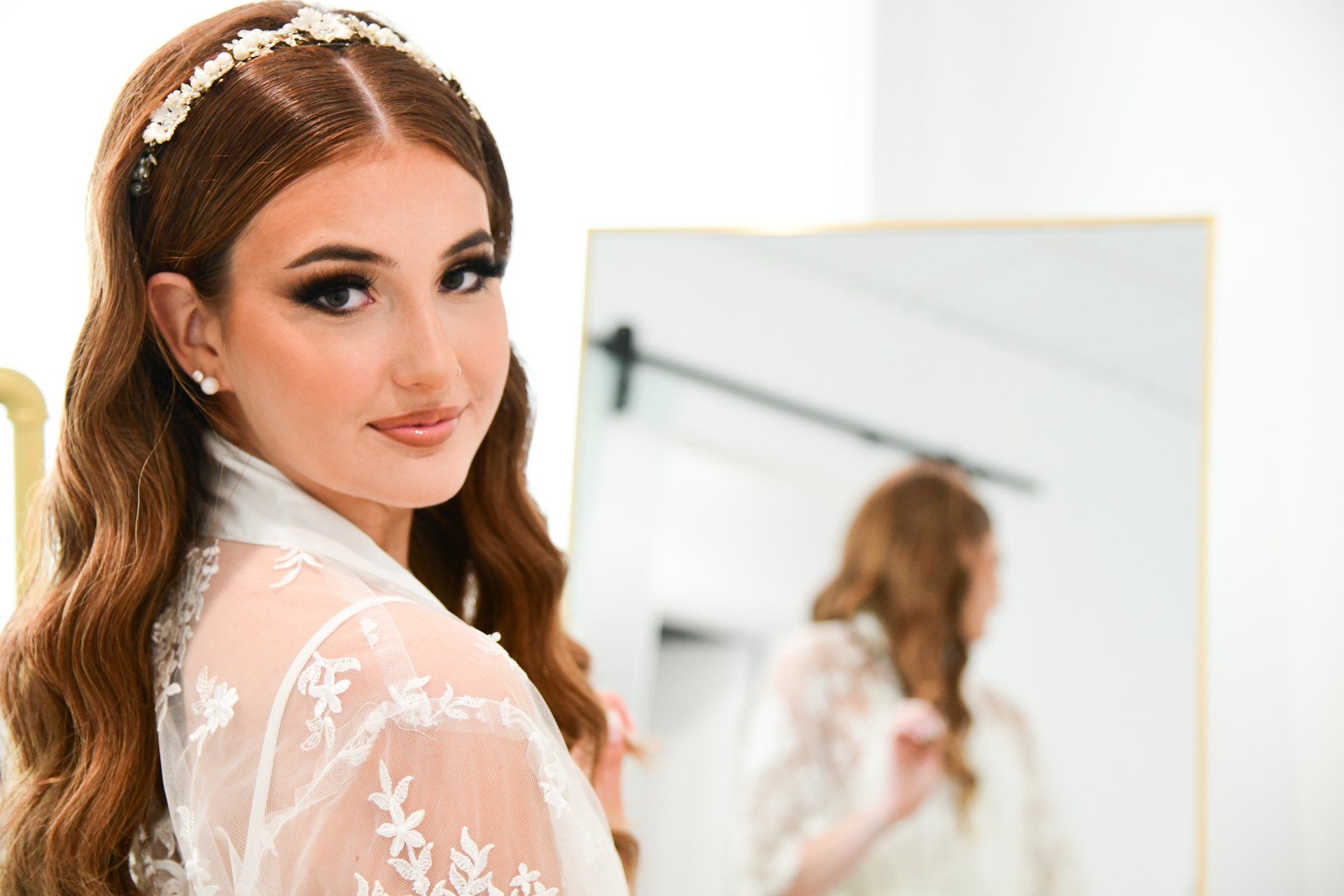 Woman with long, wavy brown hair, wearing a white floral headband and robe, looking toward the camera in front of a mirror.