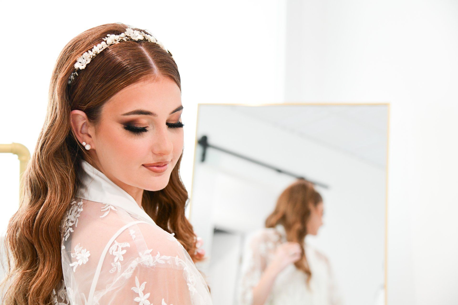 Woman with wavy red hair wearing a floral headband and lace robe, looking at a mirror.
