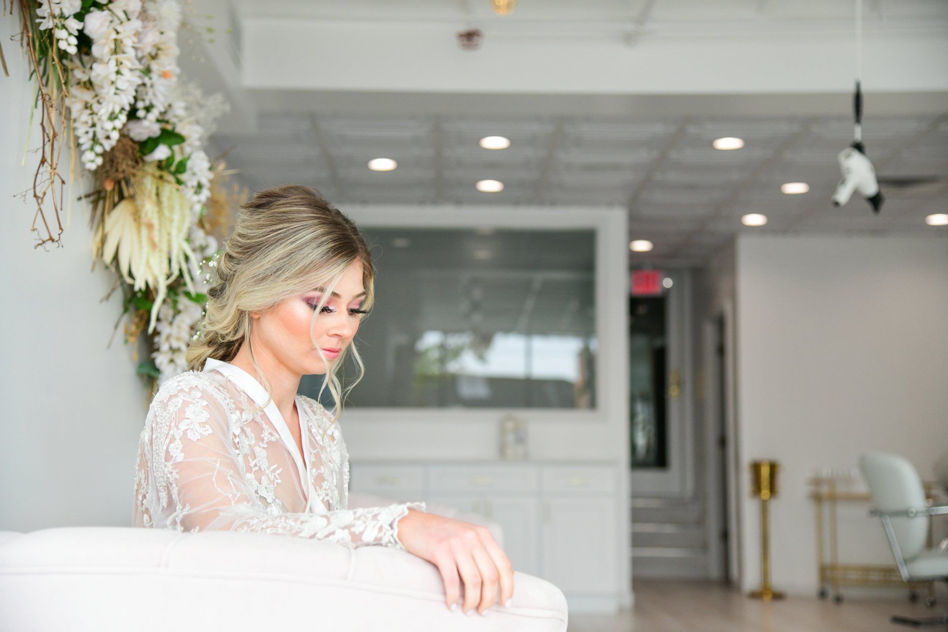 Woman in lace robe with updo hairstyle, sitting in a white room. Floral arrangement to the left.