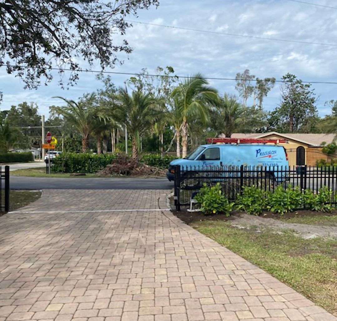 A blue service van is parked on the street behind a black metal fence, viewed from a paver driveway.